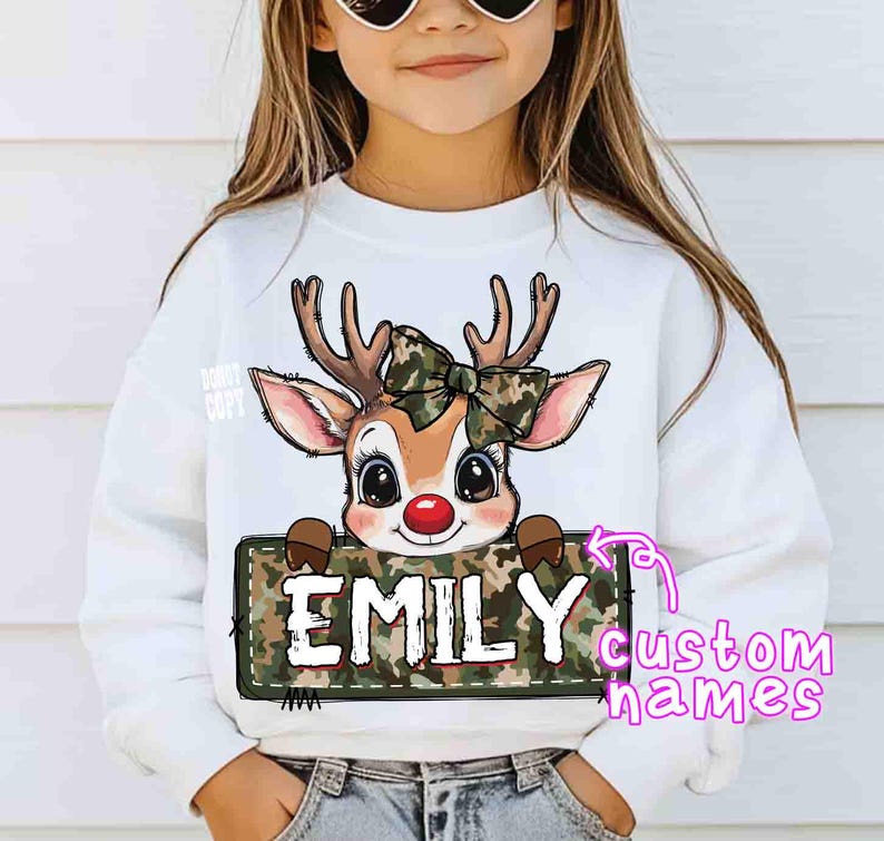 May include: White sweatshirt featuring a cartoon reindeer with a camouflage bow and a red nose. The reindeer holds a sign with the name "EMILY" in white letters. The sweatshirt is for children.