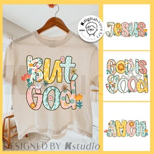 May include: A beige t-shirt with the phrase "But God" in colorful, floral-decorated letters. The design includes the words "Jesus", "God is Good", and "Amen" in a similar style. The shirt is hanging on a wooden hanger.