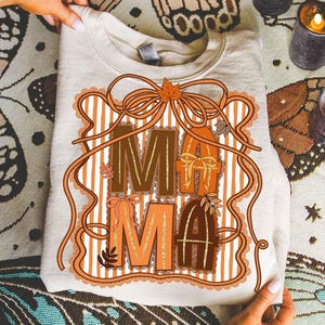 Op de afbeelding: Een beige sweatshirt met het woord "MAMA" in bruine en oranje letters. De letters zijn versierd met strikken, bladeren en een gestreepte achtergrond. Het ontwerp is omlijst met een decoratieve rand.