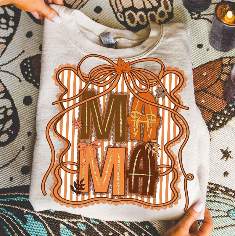 Op de afbeelding: Een beige sweatshirt met het woord "MAMA" in bruine en oranje letters. De letters zijn versierd met strikken, bladeren en een gestreepte achtergrond. Het ontwerp is omlijst met een decoratieve rand.