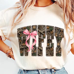 Puede incluir: Sudadera color crema con la palabra "WIFEY" en estampado de camuflaje y un lazo rosa. Las letras est&aacute;n delineadas en un color oscuro. Una prenda casual para el d&iacute;a a d&iacute;a.