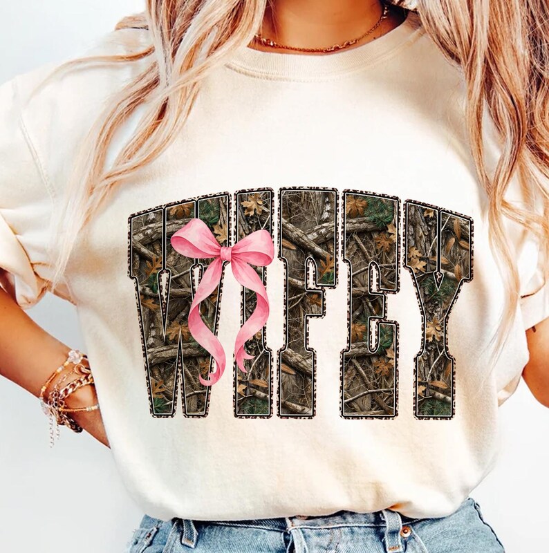 Puede incluir: Sudadera color crema con la palabra "WIFEY" en estampado de camuflaje y un lazo rosa. Las letras est&aacute;n delineadas en un color oscuro. Una prenda casual para el d&iacute;a a d&iacute;a.