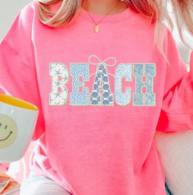 May include: A vibrant pink sweatshirt featuring the word "BEACH". The letters are crafted from patchwork fabric with patterns of starfish, polka dots, and seashells in blue and white, edged with pearls.