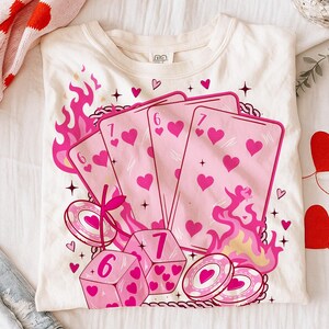 May include: Cream-colored t-shirt featuring a graphic of playing cards, dice, and casino chips with pink hearts and flames. The design includes the numbers 6 and 7 on the cards and dice, with a cherry and star accents.