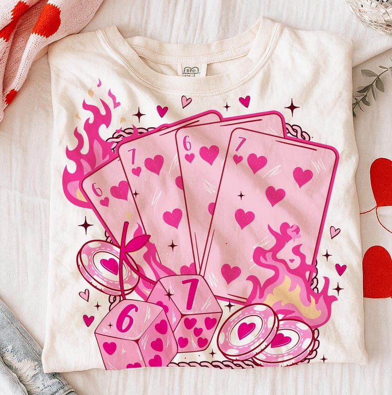 May include: Cream-colored t-shirt featuring a graphic of playing cards, dice, and casino chips with pink hearts and flames. The design includes the numbers 6 and 7 on the cards and dice, with a cherry and star accents.