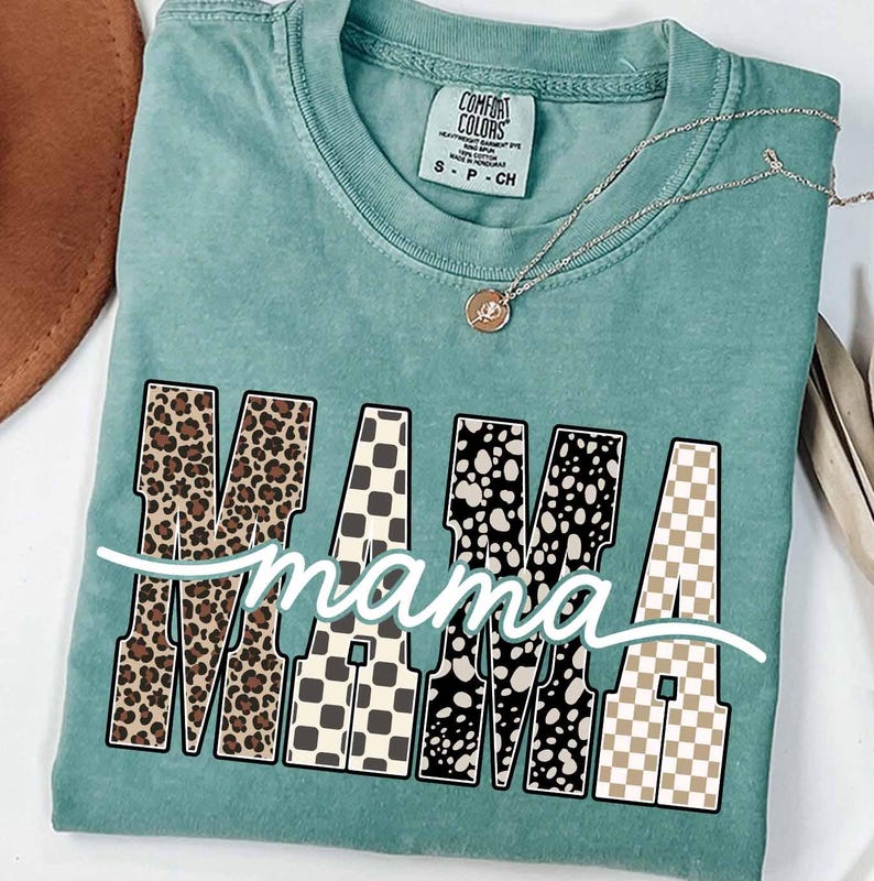 May include: A teal Comfort Colors t-shirt with the word "MAMA" in large, patterned letters. The "M"s have a leopard print, the "A"s have a checkered pattern, and the "M" in the middle has a black and white polka dot pattern. The word "mama" is written in cursive below.