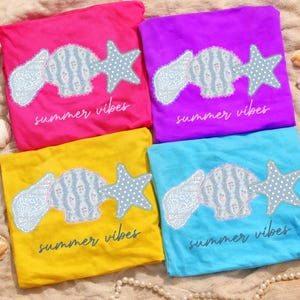 May include: Four colorful t-shirts: pink, purple, yellow, and blue. Each shirt features a seashell and starfish design with the text "summer vibes". The seashell and starfish are light blue with a white polka dot pattern.