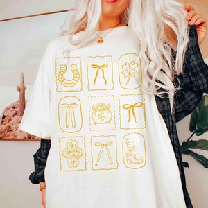 May include: White t-shirt with a gold-colored St. Patrick's Day design. The design features a horseshoe, bows, a four-leaf clover, a pot of gold, and a boot. The design is arranged in a grid pattern.