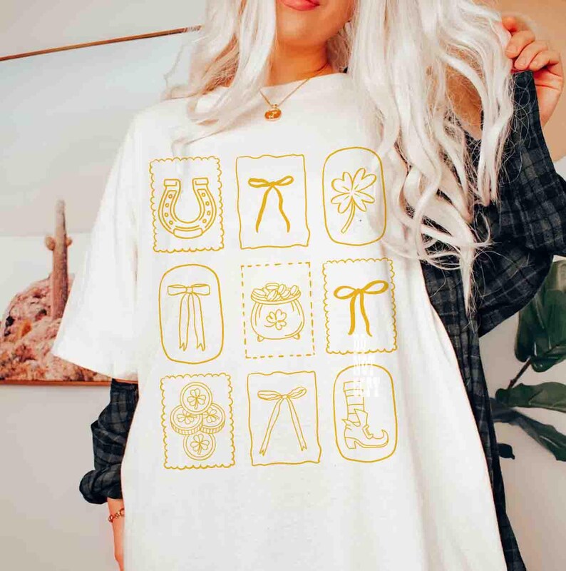 May include: White t-shirt with a gold-colored St. Patrick's Day design. The design features a horseshoe, bows, a four-leaf clover, a pot of gold, and a boot. The design is arranged in a grid pattern.