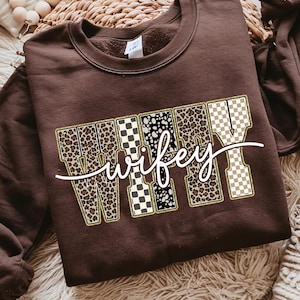 May include: A brown sweatshirt featuring the word "wifey" in a decorative font. The letters are filled with leopard print, black and white checkered patterns, and a tan and white checkered pattern. The word is outlined in gold.