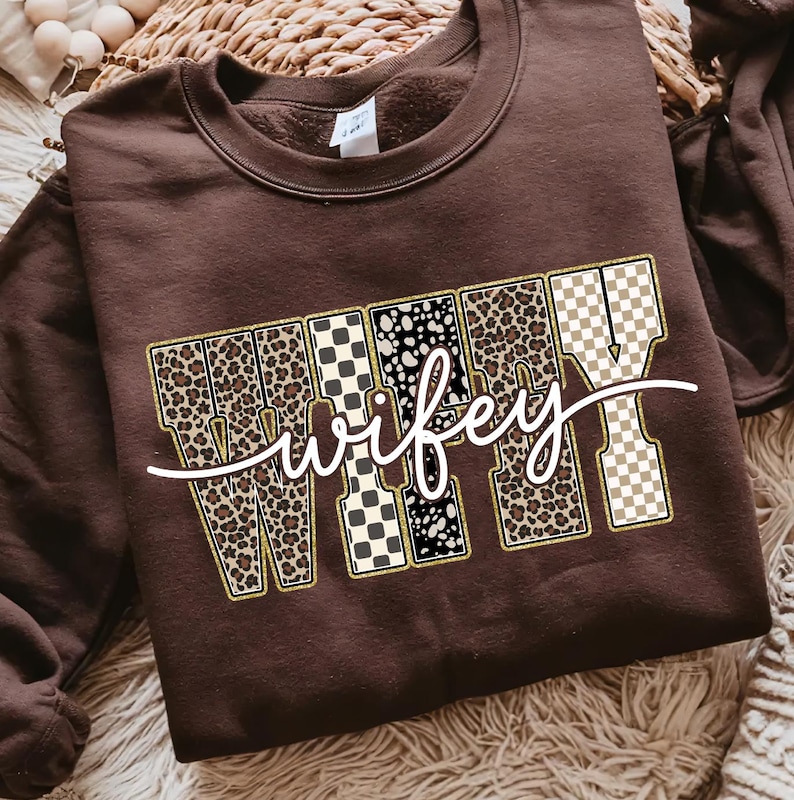 May include: A brown sweatshirt featuring the word "wifey" in a decorative font. The letters are filled with leopard print, black and white checkered patterns, and a tan and white checkered pattern. The word is outlined in gold.