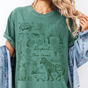 May include: Green t-shirt with a graphic of various animals including a zebra, owl, and turtle. The text "Respect the Local" is printed in a script font. The shirt is a solid green color.
