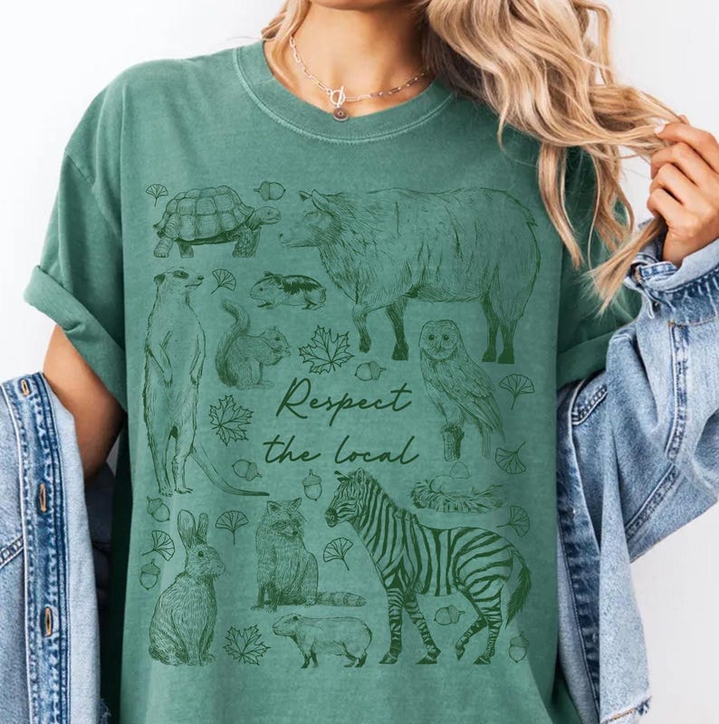 May include: Green t-shirt with a graphic of various animals including a zebra, owl, and turtle. The text "Respect the Local" is printed in a script font. The shirt is a solid green color.
