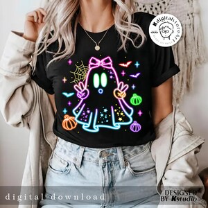 May include: Black t-shirt featuring a neon ghost design with peace signs, pumpkins, bats, and stars. The ghost has a bow and is surrounded by a spiderweb. Text on the shirt reads "digital download" and "designed by Kstudio".