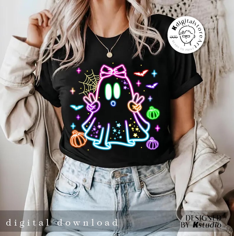 May include: Black t-shirt featuring a neon ghost design with peace signs, pumpkins, bats, and stars. The ghost has a bow and is surrounded by a spiderweb. Text on the shirt reads "digital download" and "designed by Kstudio".