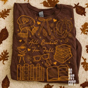 May include: Brown long-sleeve t-shirt with a fall-themed graphic. The design includes a scarf, pumpkin, books, glasses, and the text "All Booked For Fall." The shirt has a crew neck and is folded.