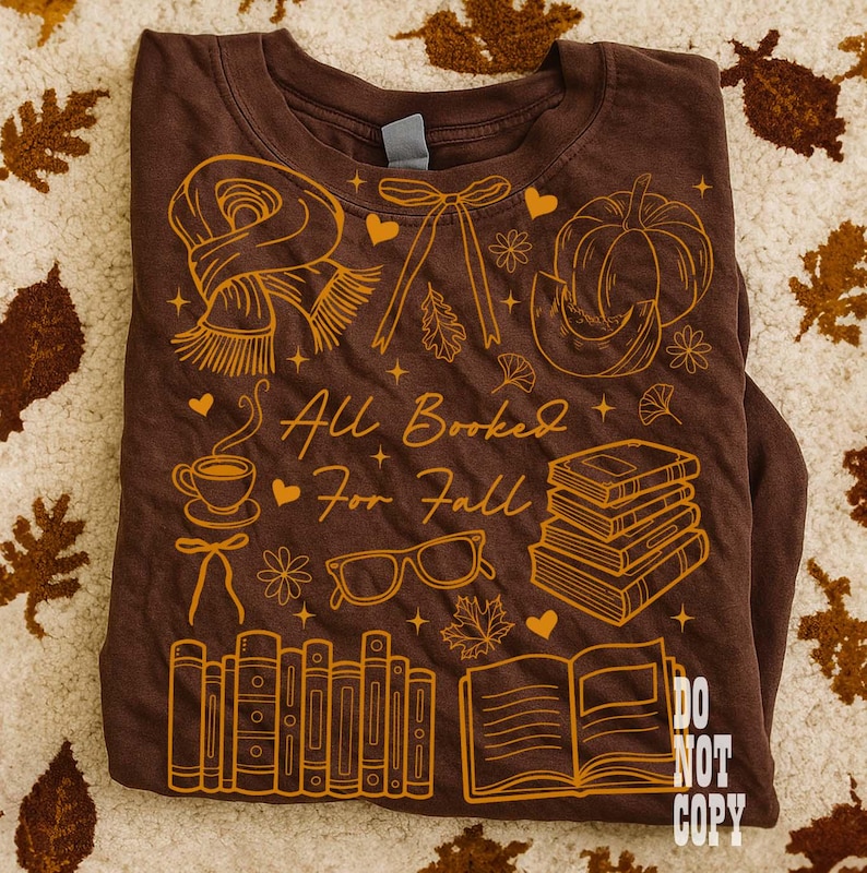 May include: Brown long-sleeve t-shirt with a fall-themed graphic. The design includes a scarf, pumpkin, books, glasses, and the text "All Booked For Fall." The shirt has a crew neck and is folded.