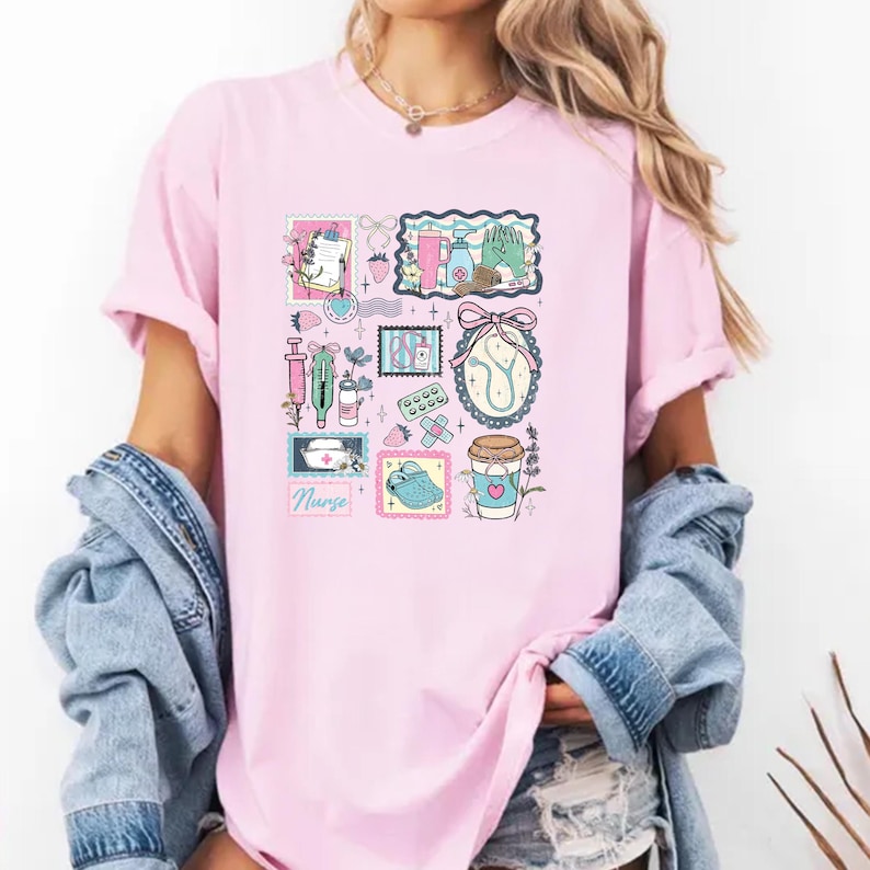 Op de afbeelding: Zachtroze T-shirt met een kleurrijke collage met een medisch thema. Het ontwerp bevat een spuit, pillen en een stethoscoop. Het woord "Nurse" staat onderaan gedrukt. Het shirt heeft korte mouwen en een relaxte pasvorm.