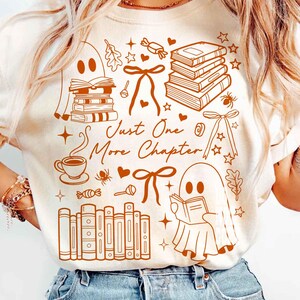 May include: Cream-colored t-shirt with a brown graphic design featuring books, ghosts, bows, and the text "Just One More Chapter." The design includes a coffee cup, stars, and candy, creating a cozy, book-themed aesthetic.