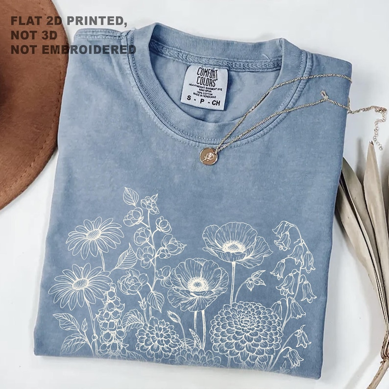 May include: A blue Comfort Colors t-shirt with a white floral design. The shirt has a crew neck and short sleeves. A gold necklace with a pendant is draped across the shirt. The text "FLAT 2D PRINTED, NOT 3D, NOT EMBROIDERED" is in the upper left corner.