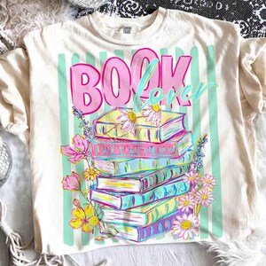 May include: Cream-colored t-shirt with a colorful graphic. The design features a stack of books with floral accents and the words "BOOK Lover" in pink and light blue. Vertical mint green stripes are behind the text.