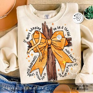 May include: A beige sweatshirt featuring a brown wooden cross with an orange ribbon. The text "If you're hanging on by a thread make sure it's the hem of His garment" is printed on the sweatshirt.