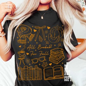 May include: Black t-shirt with a gold graphic design featuring fall-themed illustrations, including a scarf, pumpkin, books, and the text "All Booked For Fall". The design also includes hearts, leaves, and a coffee cup.