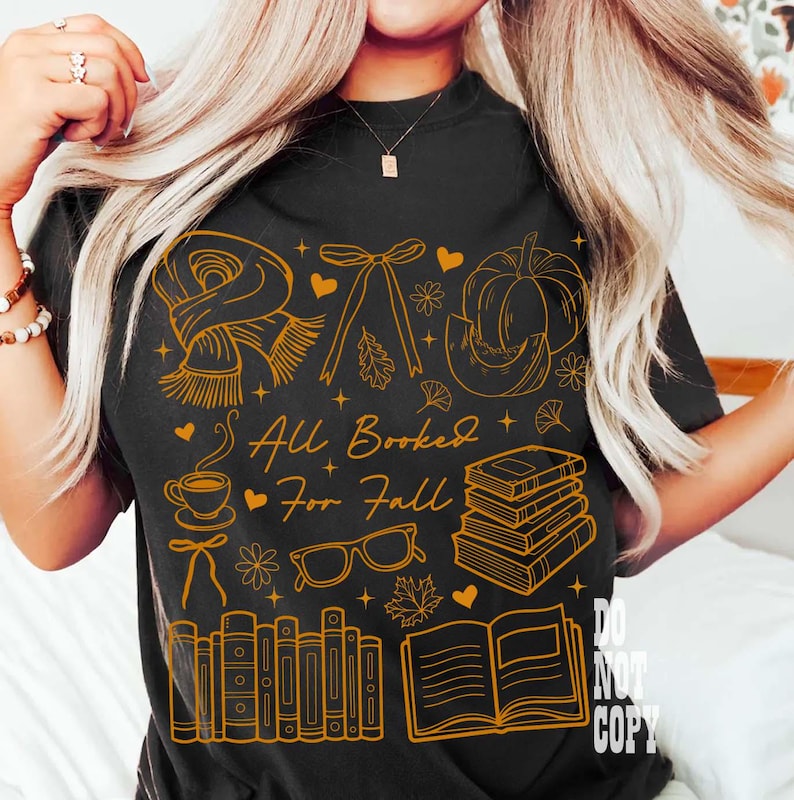 May include: Black t-shirt with a gold graphic design featuring fall-themed illustrations, including a scarf, pumpkin, books, and the text "All Booked For Fall". The design also includes hearts, leaves, and a coffee cup.