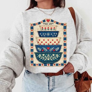 Pu&ograve; includere: Una felpa grigio chiaro con un design a ciotole impilate. Il design include quattro ciotole nei toni del blu e del crema, decorate con cuori, fiori e motivi a foglie. Il design &egrave; incorniciato da un bordo blu e accenti di cuori rossi.