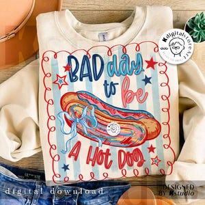 May include: Cream-colored sweatshirt with a graphic of a hot dog and the text "BAD day to be A Hot Dog." The design includes red, white, and blue elements, with a decorative border and stars. The sweatshirt is a digital download.