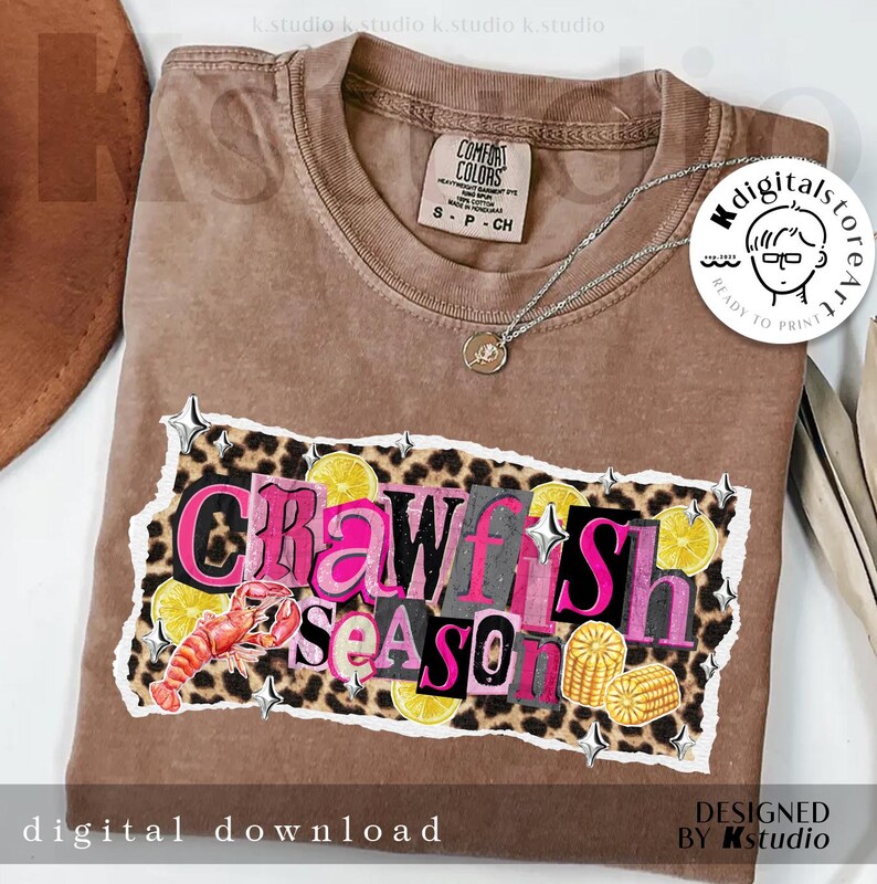 May include: Brown Comfort Colors t-shirt with a graphic that says "Crawfish Season" in pink and gray letters. The design includes a crawfish, lemons, and corn on a leopard print background. A silver necklace is draped on the shirt.