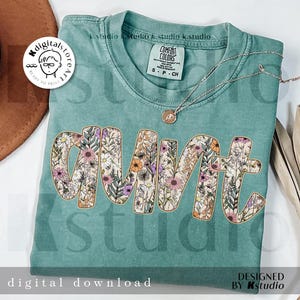 May include: A teal t-shirt with a floral design that spells out "aunt". The design is in a light brown outline with a variety of flowers and leaves in shades of pink, purple, yellow, and green.