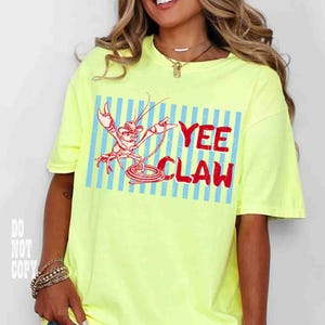 May include: A neon yellow t-shirt with a red and blue graphic. The graphic features a cartoon crawfish wearing a cowboy hat and holding a rope, with the words "YEE CLAW" in red. The shirt has short sleeves.