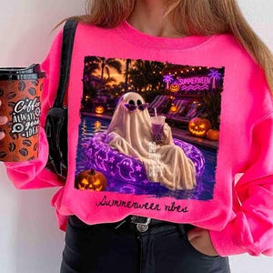 May include: A bright pink sweatshirt featuring a graphic of a ghost in sunglasses relaxing in a pool with a drink, surrounded by pumpkins and the text "Summerween vibes". A coffee cup is held in the left hand.