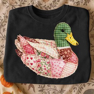 May include: Black sweatshirt featuring a patchwork duck design. The duck has a green checkered head, a yellow beak, and wings and body made of various floral and patterned fabrics in pinks, reds, and whites. The design is centered on the chest.