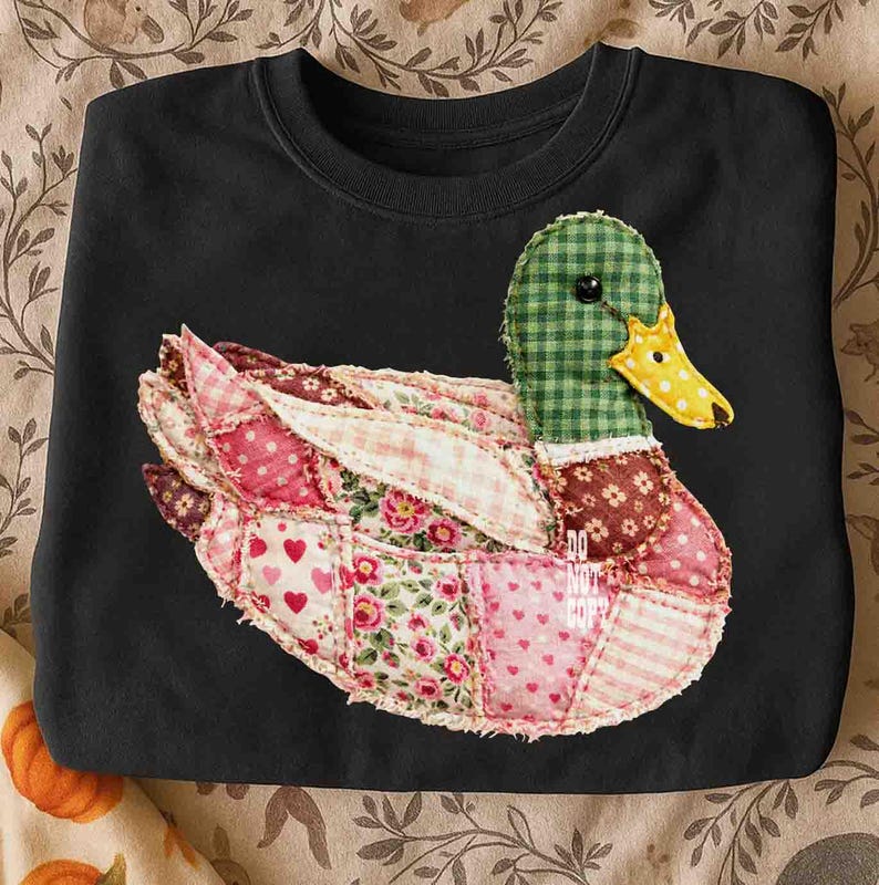 May include: Black sweatshirt featuring a patchwork duck design. The duck has a green checkered head, a yellow beak, and wings and body made of various floral and patterned fabrics in pinks, reds, and whites. The design is centered on the chest.