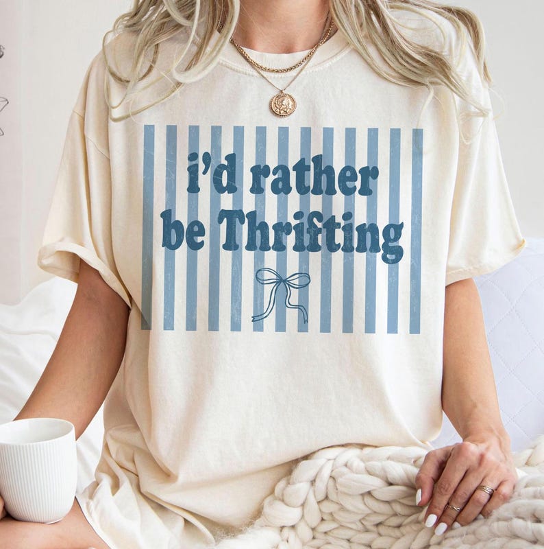 May include: Cream-colored t-shirt with the text "I'd rather be Thrifting" in blue, set against a backdrop of blue vertical stripes. A small bow graphic is below the text. The shirt is worn by a person.