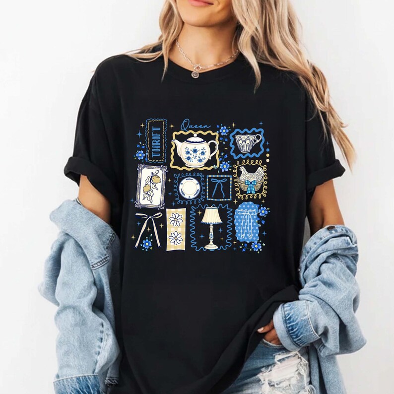 May include: Black t-shirt featuring a collage of vintage-style illustrations in blue and yellow. The design includes a teapot, teacup, lemon sprig, plate, bow, chicken, lamp, and a blue patterned curtain. The word "THRIFT" is printed vertically.