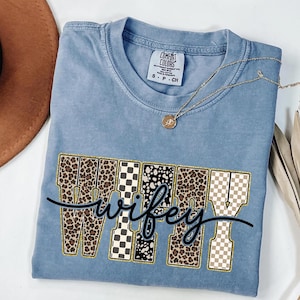 May include: A blue Comfort Colors t-shirt featuring the word "wifey" in a decorative font. The letters are filled with leopard print, black and white patterns, and a gold outline. A gold necklace with a pendant is draped on the shirt.