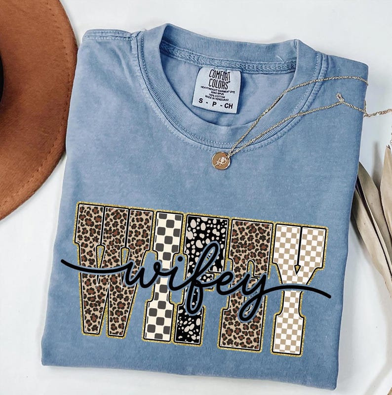 May include: A blue Comfort Colors t-shirt featuring the word "wifey" in a decorative font. The letters are filled with leopard print, black and white patterns, and a gold outline. A gold necklace with a pendant is draped on the shirt.