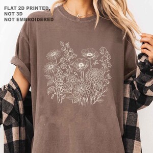 May include: A brown t-shirt featuring a white floral design. The design includes various flowers and foliage. The text "FLAT 2D PRINTED, NOT 3D, NOT EMBROIDERED" is printed in the upper left corner. The shirt has rolled-up sleeves.