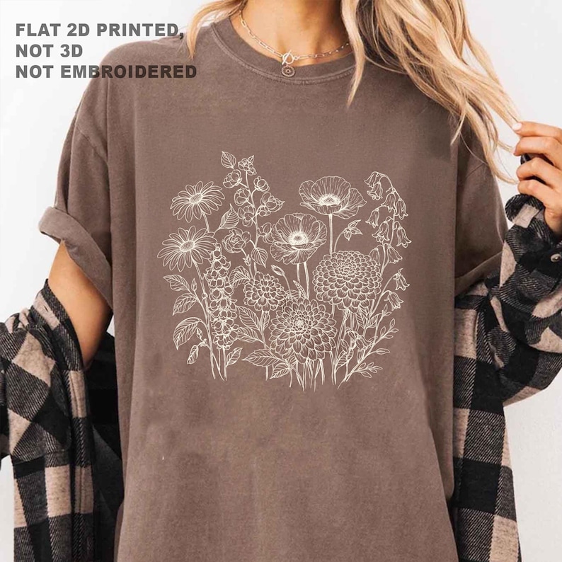 May include: A brown t-shirt featuring a white floral design. The design includes various flowers and foliage. The text "FLAT 2D PRINTED, NOT 3D, NOT EMBROIDERED" is printed in the upper left corner. The shirt has rolled-up sleeves.