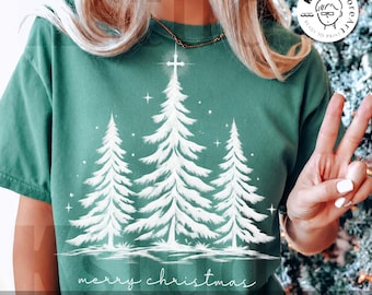 May include: A forest green t-shirt featuring a white graphic of three pine trees with a cross at the top of the centre tree. The design includes small white stars and the text "merry christmas" in a cursive script at the bottom of the graphic.