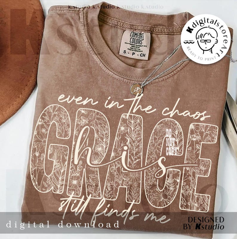 May include: A brown Comfort Colors t-shirt with the words "even in the chaos GRACE still finds me" in white script and floral design. A silver necklace with a pendant is on the shirt. The shirt is a digital download.