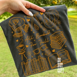 May include: Dark gray t-shirt with an orange graphic design featuring fall-themed illustrations like a scarf, pumpkin, books, and glasses. The text "All Booked For Fall" is also included in the design.
