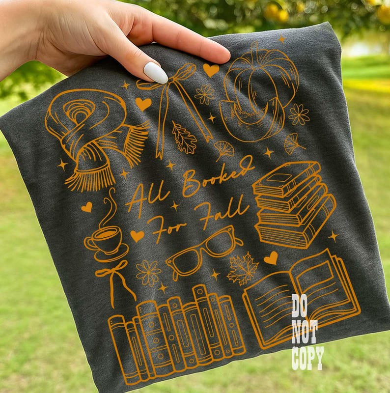 May include: Dark gray t-shirt with an orange graphic design featuring fall-themed illustrations like a scarf, pumpkin, books, and glasses. The text "All Booked For Fall" is also included in the design.