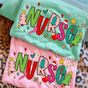May include: Two Christmas-themed t-shirts, one mint green and one pink, with the word "NURSE" in large, colorful letters. The design includes Christmas trees, ornaments, candy canes, and a gingerbread man. The shirts are folded.