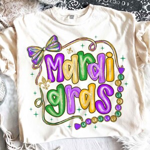 May include: Cream-colored t-shirt featuring the words "Mardi Gras" in large, colorful letters. The design includes a purple and green bow, a strand of beads, and gold accents. The shirt is a Comfort Colors brand.