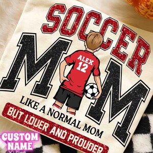 May include: Cream-colored t-shirt with a soccer theme. The design features the words "SOCCER MOM" in red glitter and black letters. A cartoon figure in a red jersey with the name "ALEX" and the number "12" is holding a soccer ball. The text "LIKE A NORMAL MOM BUT LOUER AND PROUDER" is also included.