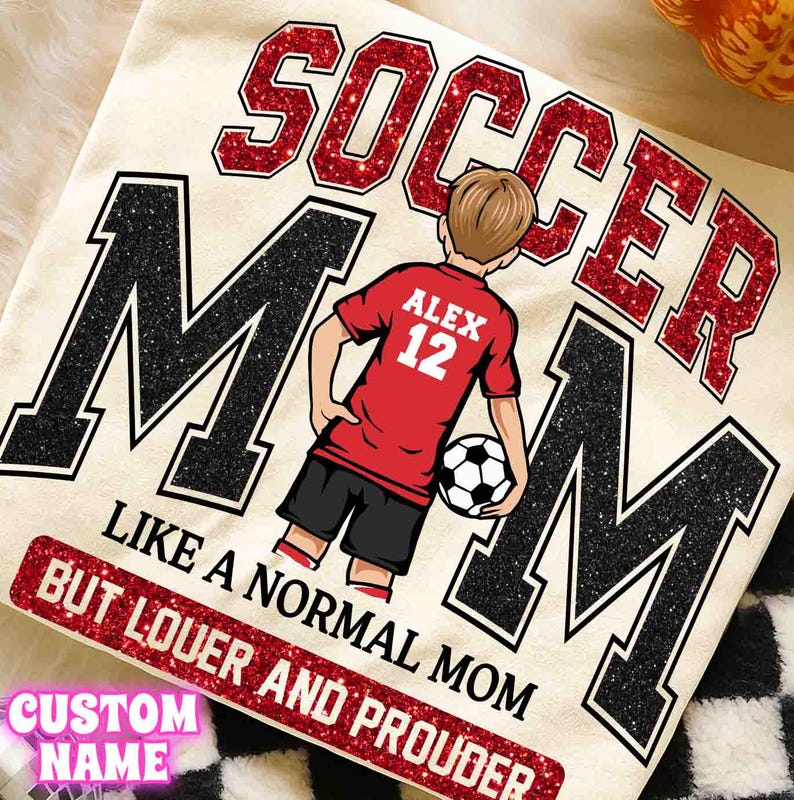 May include: Cream-colored t-shirt with a soccer theme. The design features the words "SOCCER MOM" in red glitter and black letters. A cartoon figure in a red jersey with the name "ALEX" and the number "12" is holding a soccer ball. The text "LIKE A NORMAL MOM BUT LOUER AND PROUDER" is also included.